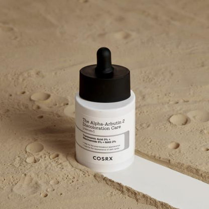 The COSRX The Alpha-Arbutin 2 Discoloration Care Serum 50ml with a dropper cap sits on a textured sandy surface, highlighting ingredients such as niacinamide, mandelic acid, and tranexamic acid for improved skin clarity.