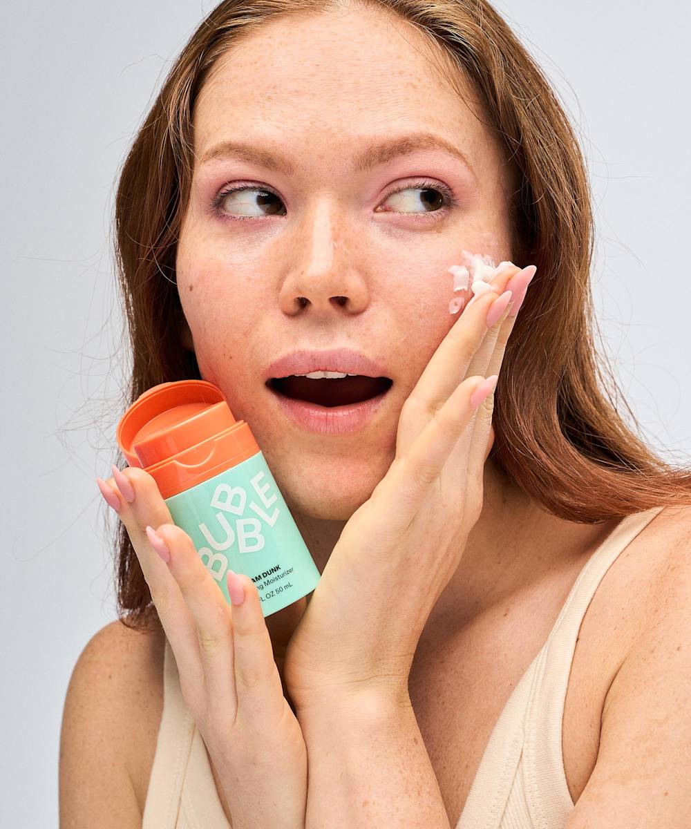 Someone with long hair applies Bubble Slam Dunk Hydrating Moisturiser Cream from a turquoise container to their cheek. The orange cap highlights their surprised or delighted expression as the Aloe Leaf Juice-rich formula works wonders.