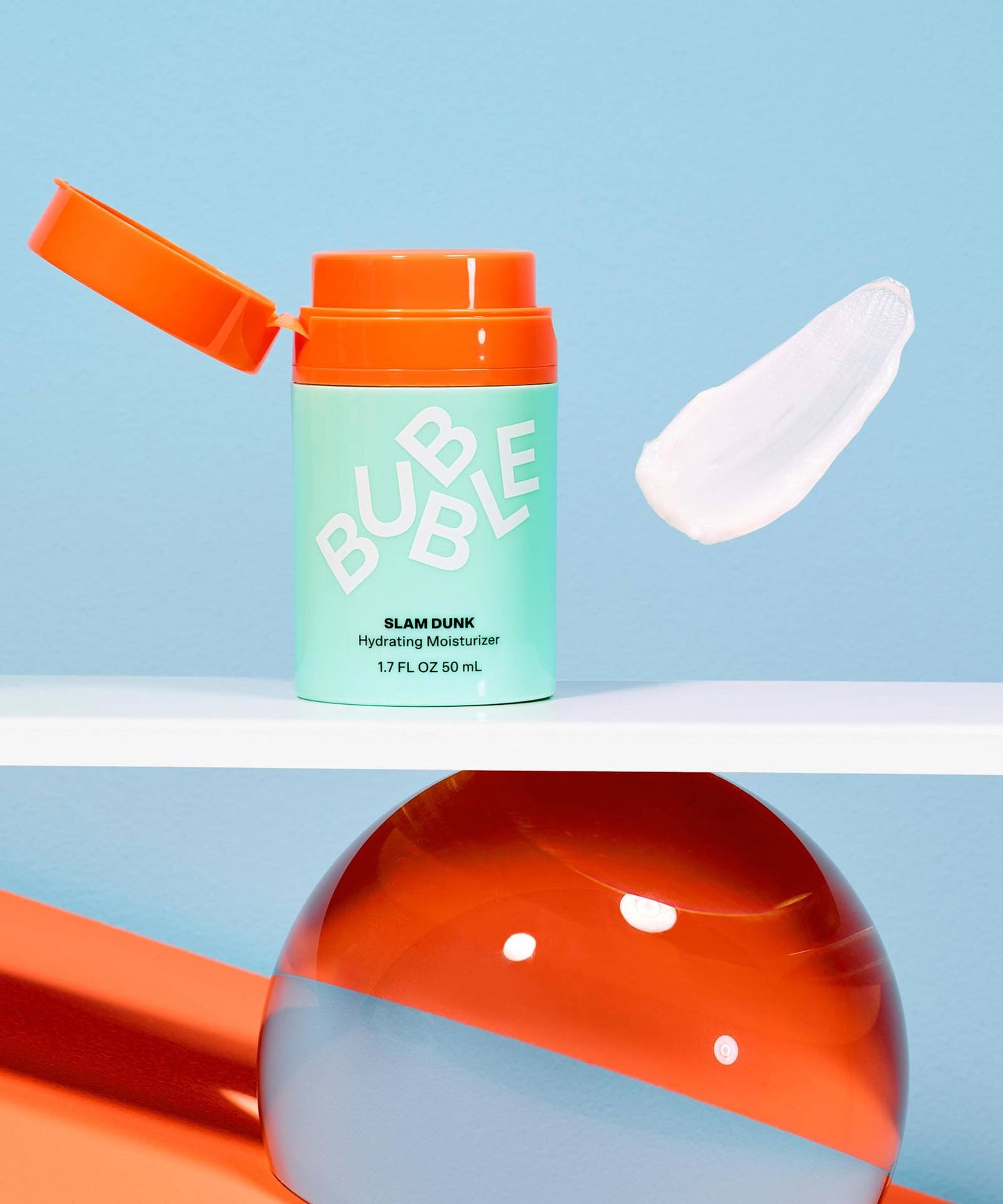 The image displays a vibrant skincare product: an open mint-green bottle labeled Bubble Slam Dunk Hydrating Moisturiser Cream 50ml with an orange cap beside a smear of cream. It rests on a clear sphere and orange surface, all against a blue background.