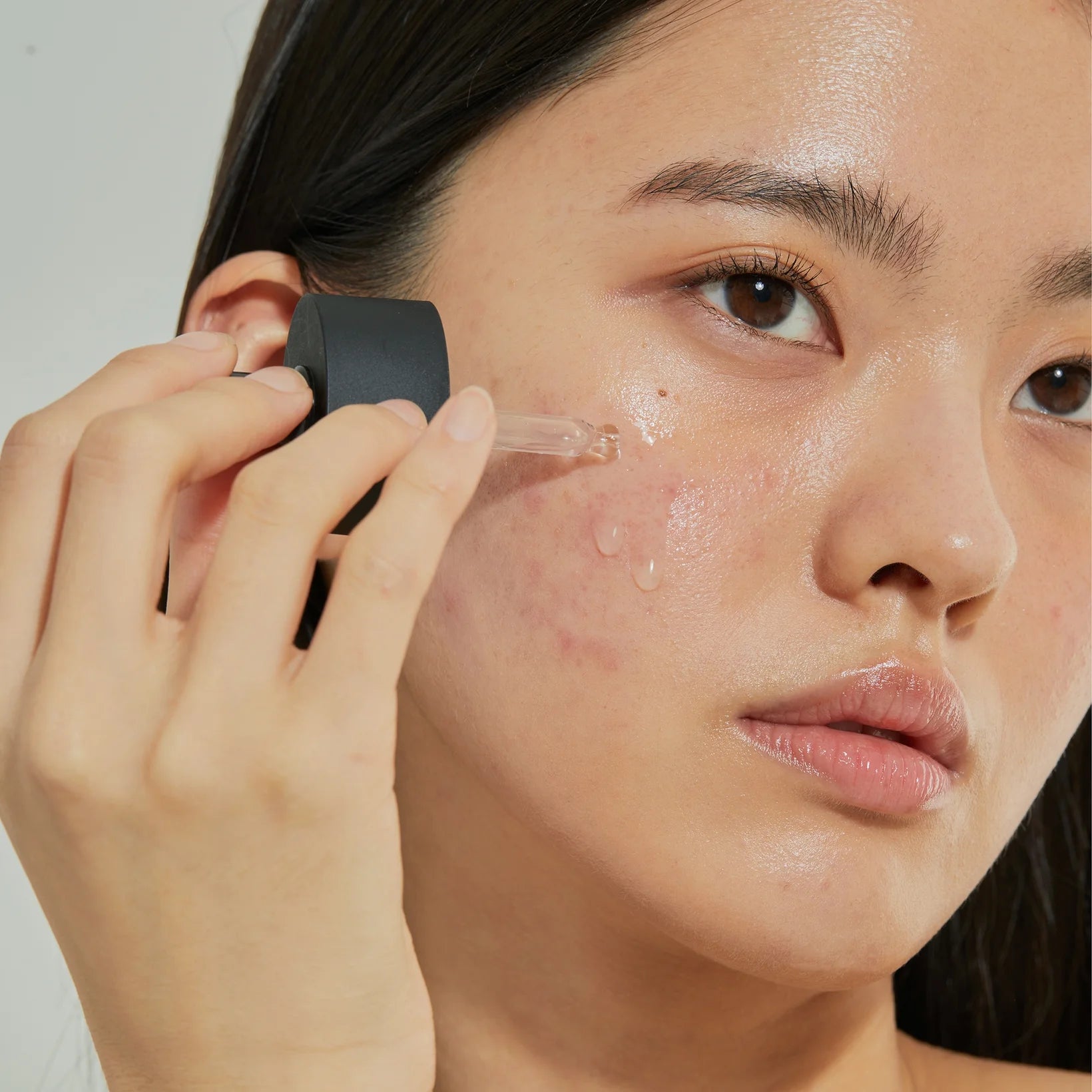 A person applies COSRX The Hyaluronic Acid 3 Serum (20ml) with a dropper to their cheek, revealing clear skin with visible pores and some redness. The serum’s amino acids and Ceramide NP help plump skin and support its moisture barrier.