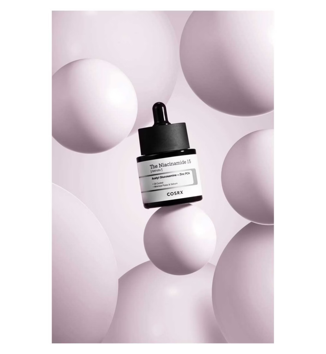 A COSRX The Niacinamide 15 Serum – 20ml bottle with dropper floats among soft pink spheres on a matching background, showcasing its 15% niacinamide formula to reduce oil and fight acne.
