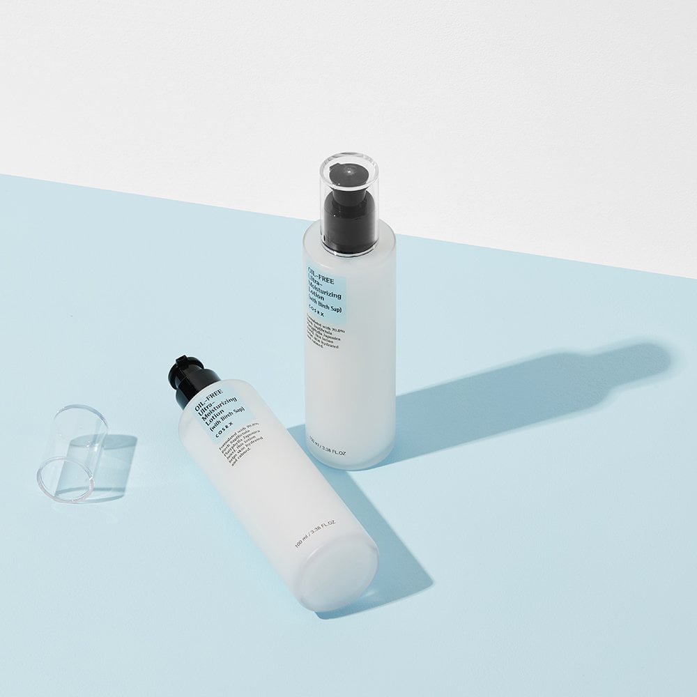 Against a light blue and white backdrop, two cylindrical COSRX bottles featuring Korean text showcase clean beauty. One stands vertically while the other lies horizontally. Transparent caps cast subtle shadows, with the COSRX Oil-Free Ultra Moisturizing Lotion promising radiant skin through nourishing birch sap.