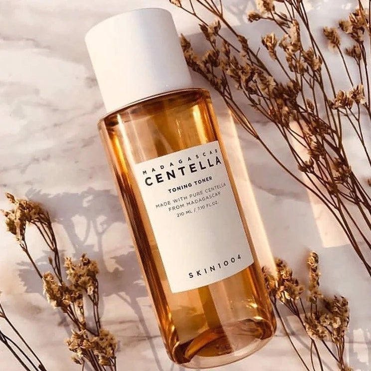 A bottle of SKIN1004 Madagascar Centella Toning Toner placed on a marble surface, surrounded by dried flowers. The transparent bottle contains a golden-hued liquid, and it has a white screw cap. The label displays the product name and details, emphasizing that it is made with pure Centella from Madagascar. The warm lighting and natural elements create an aesthetic and calming presentation.