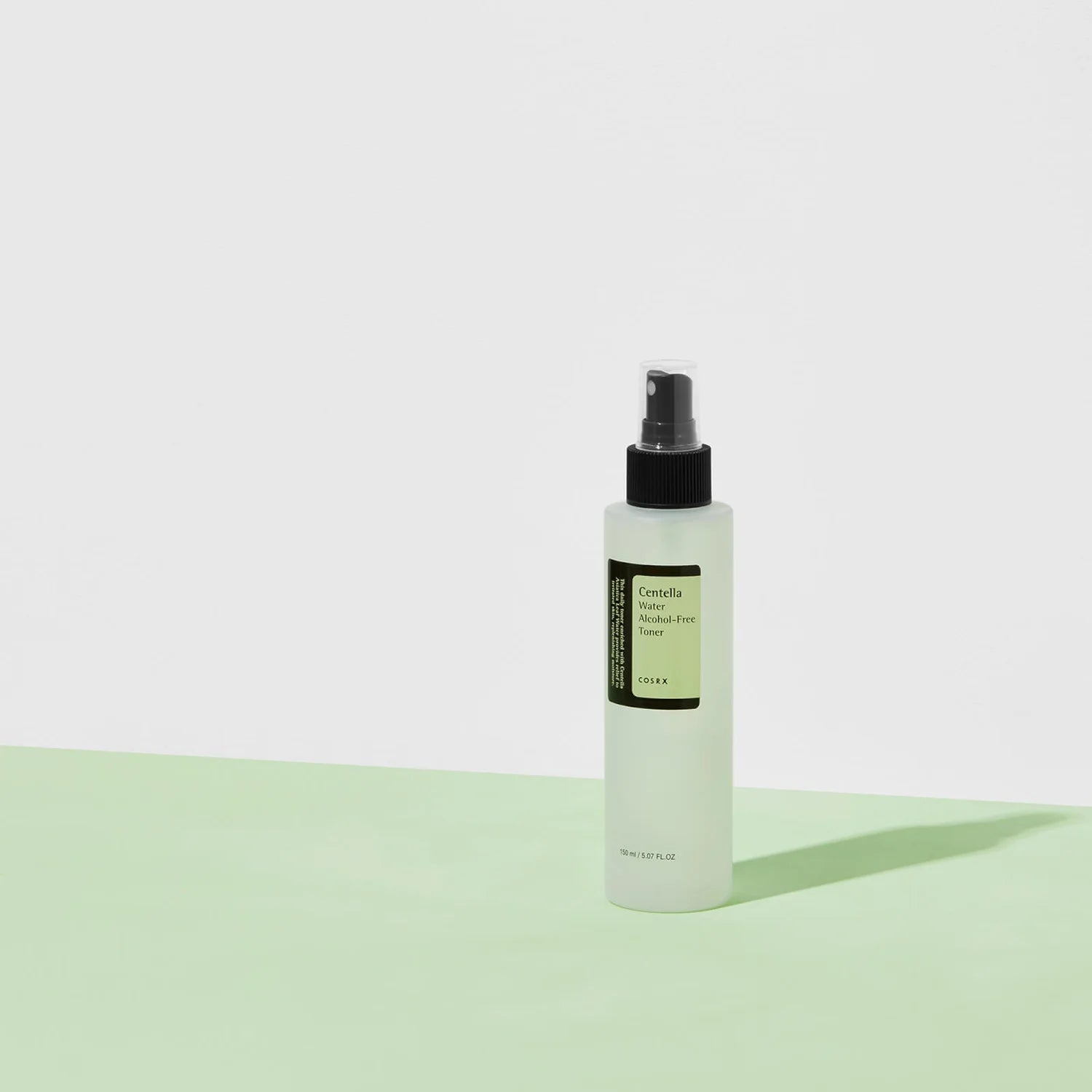 A translucent spray bottle of COSRX Centella Water Alcohol-Free Toner with a black nozzle stands on a light green surface. The minimalist green and white label highlights its soothing properties. Shadows are elegantly cast onto the white background beneath it.