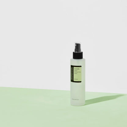 A translucent spray bottle of COSRX Centella Water Alcohol-Free Toner with a black nozzle stands on a light green surface. The minimalist green and white label highlights its soothing properties. Shadows are elegantly cast onto the white background beneath it.