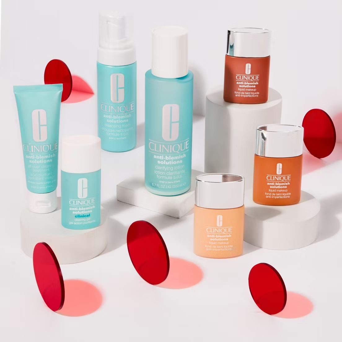 A Clinique display features Anti-Blemish Solutions Clarifying Lotion 200ml in blue and orange bottles and jars, arranged on white podiums with red transparent discs scattered around them.