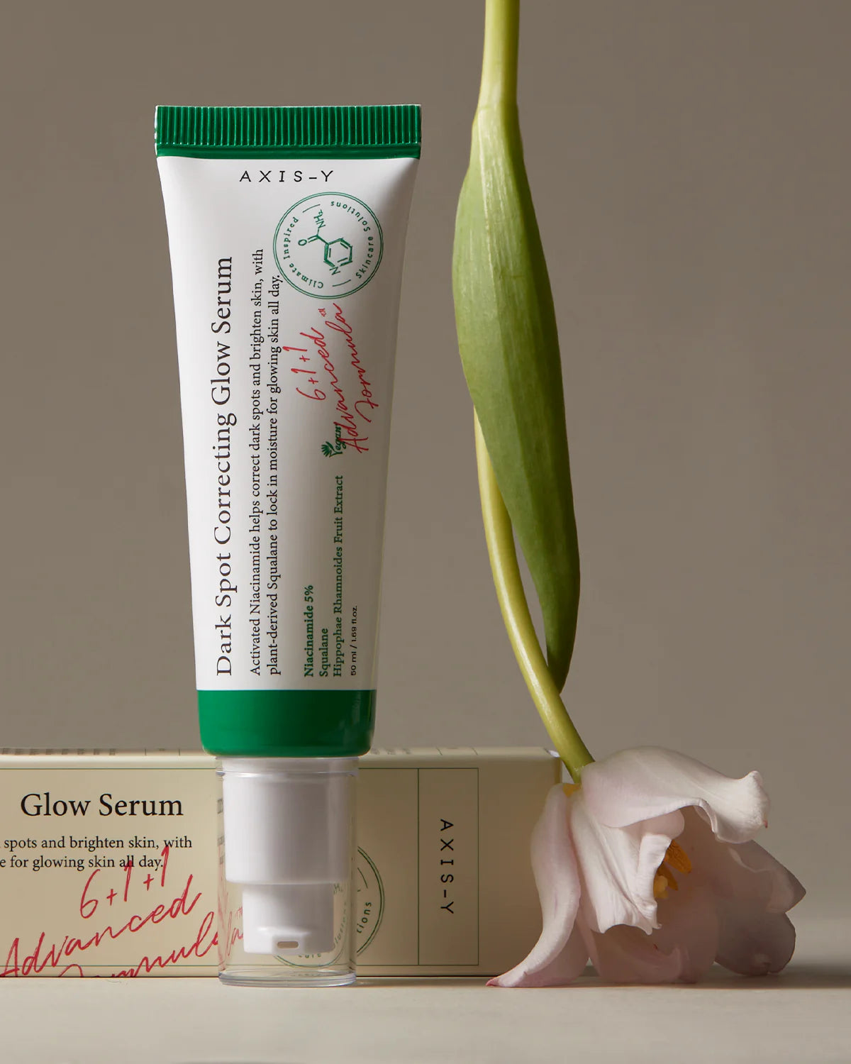 The AXIS-Y Dark Spot Correcting Glow Serum 50ml, highlighted by its glow-enhancing green and white packaging, stands upright supported by a white and pink tulip against a neutral backdrop, showcasing its niacinamide-infused benefits.
