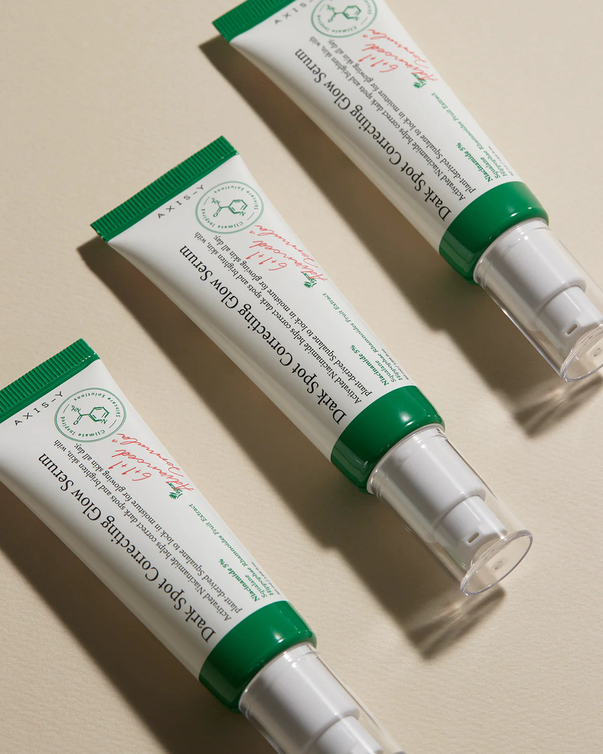 Three tubes of AXIS-Y Dark Spot Correcting Glow Serum 50ml are arranged in a diagonal pattern on a beige surface. Each is enriched with niacinamide, features a green cap, clear pump dispenser, and has a white label with red and green text.