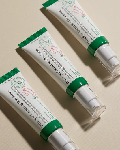 Three tubes of AXIS-Y Dark Spot Correcting Glow Serum 50ml are arranged in a diagonal pattern on a beige surface. Each is enriched with niacinamide, features a green cap, clear pump dispenser, and has a white label with red and green text.