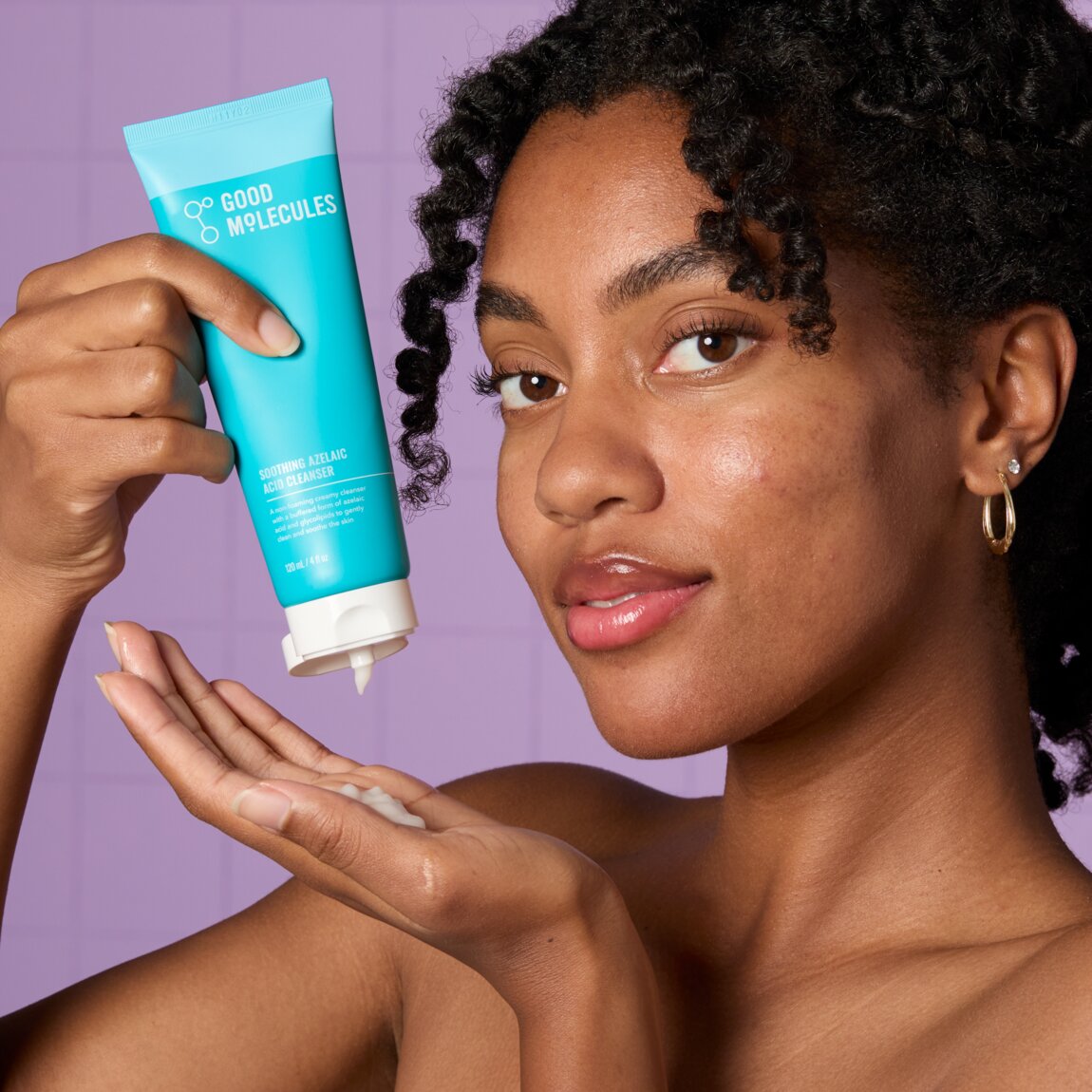 Woman applying cream from a 'Good Molecules' tube against a purple background