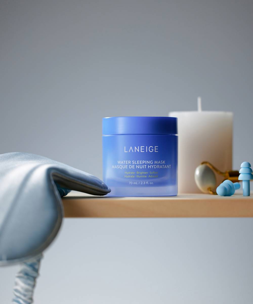 A Laneige Water Sleeping Mask - 70ml jar relaxes on a wooden surface beside a white candle, golden spoon, silk sleep mask, and blue earplugs. The softly lit backdrop sets the stage for an overnight hydrating ritual with serenity and calm provided by LANEIGE.