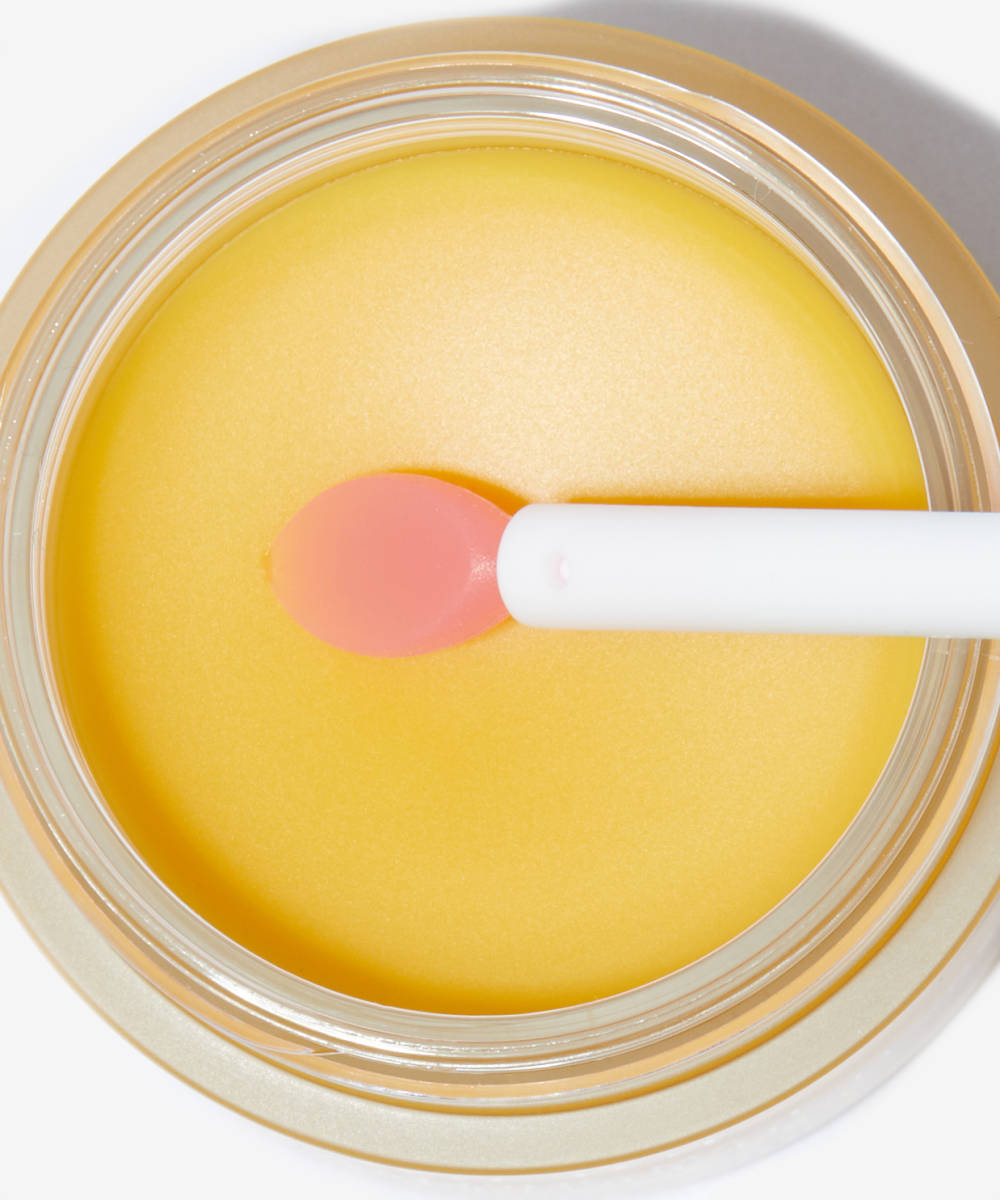 A close-up shows a round container with smooth yellow balm similar to the Laneige Lip Sleeping Mask - Mango 20g, by LANEIGE. A white-handled applicator with a pink tip rests on it, and the container has a transparent and gold lid.