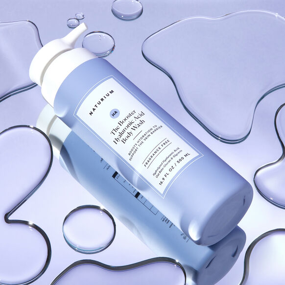 A light purple bottle of Naturium The Booster Hyaluronic Acid Body Wash 500ml with a white pump and rectangular front label rests on a shiny surface surrounded by water puddles.