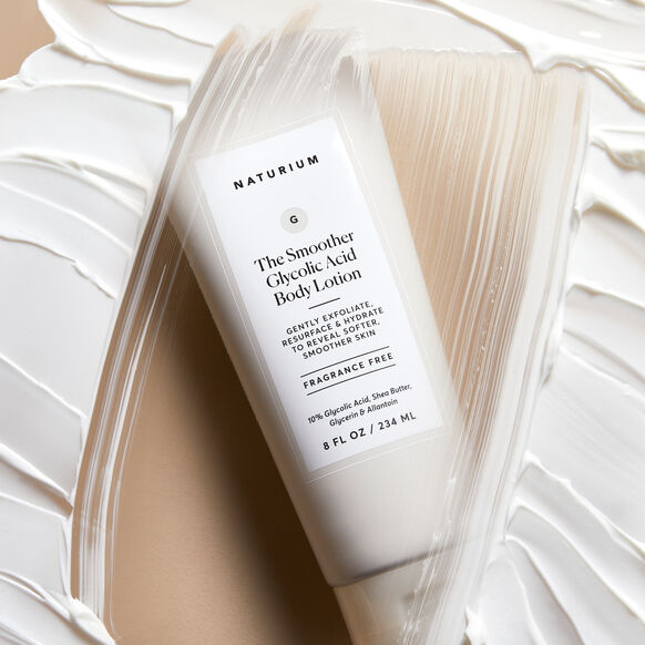 A white tube of NATURIUM The Smoother Glycolic Acid Body Lotion (234ml) by Naturium rests on a textured surface with lotion swirls, showing its label and fragrance-free formula.
