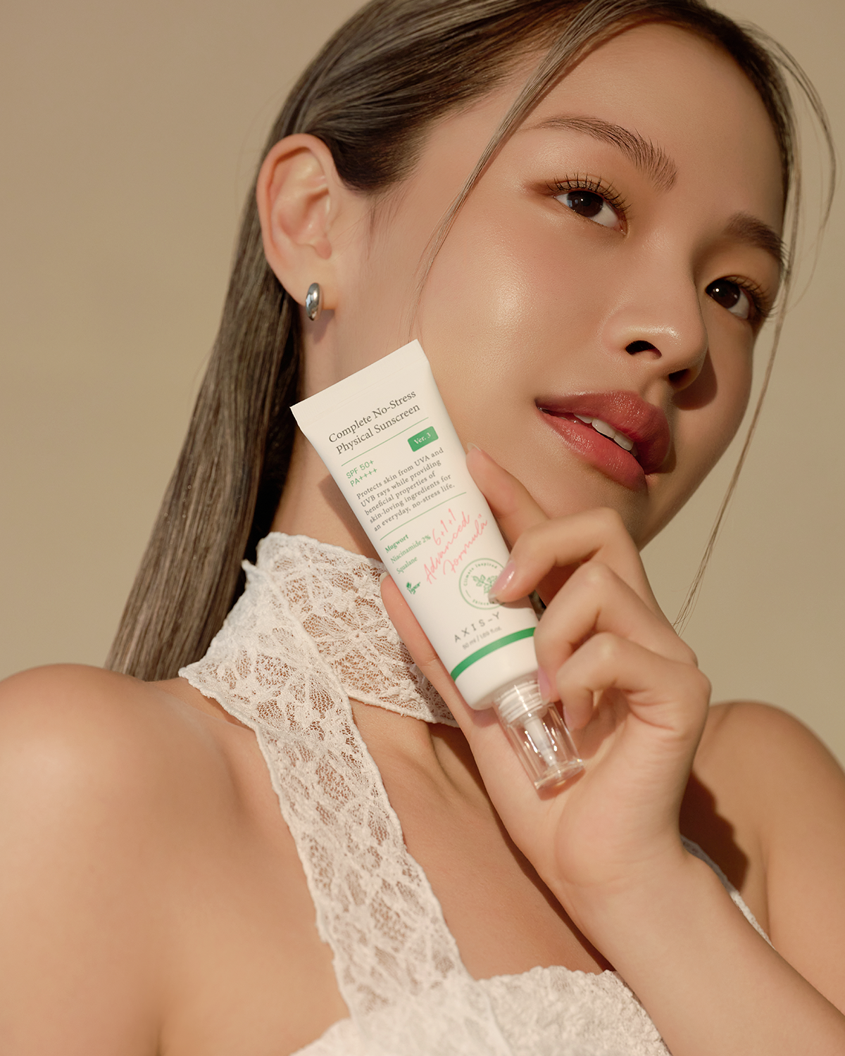 A person with long hair gently holds a tube of Axis-Y Complete No-Stress Physical Sunscreen 50ml by AXIS - Y near their face. They wear a white lace top, display a gentle expression, and stand against a soft, neutral background.