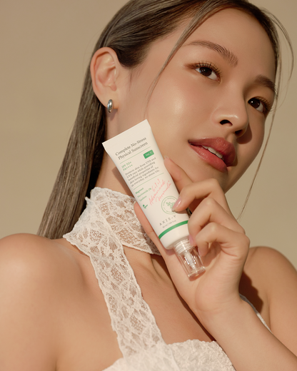 A person with long hair gently holds a tube of Axis-Y Complete No-Stress Physical Sunscreen 50ml by AXIS - Y near their face. They wear a white lace top, display a gentle expression, and stand against a soft, neutral background.