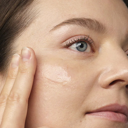 Using The Ordinary Niacinamide 10% + Zinc 1% serum, a person applies it to their cheek with two fingers, highlighting their smooth skin and blue eyes. Soft lighting enhances the complexion, making it perfect for blemish-prone skin seeking radiance.