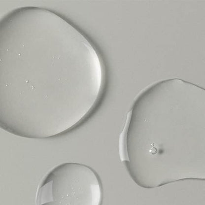 Clear liquid droplets on a smooth, light grey surface capture the essence of Abib Pore Cleansing Oil Heartleaf Oil-Wash. They vary in size and shape, contrasting the matte background with their glossy appearance, evoking a refreshing pore-clearing experience.