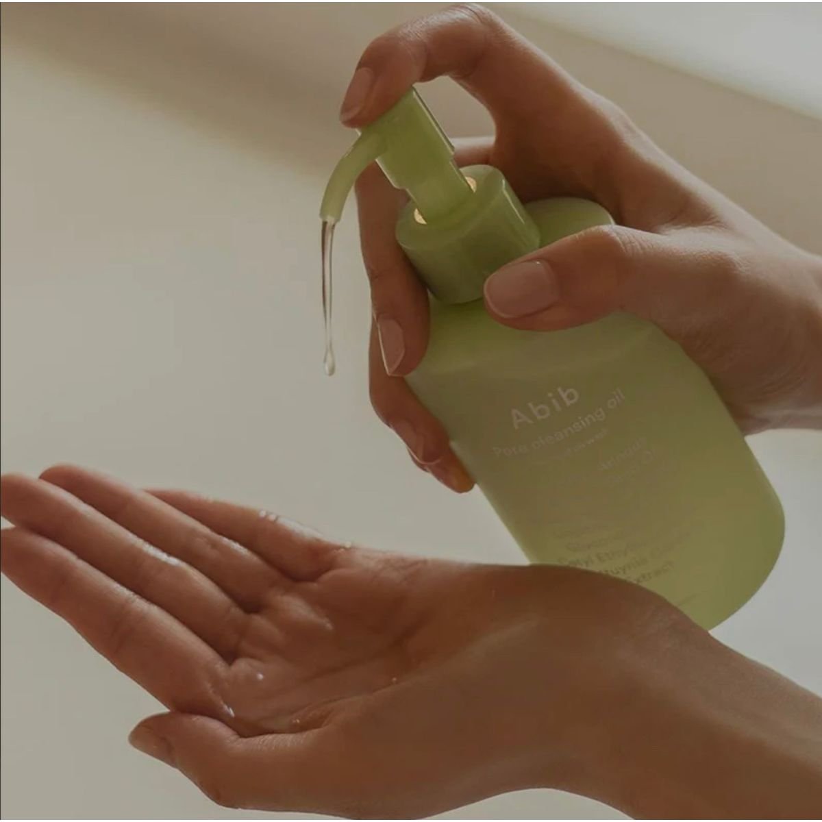 A person dispenses Abib Pore Cleansing Oil Heartleaf Oil-Wash 200ml from a light green pump bottle into their palm, with the bottle set against a soft, neutral background lit by sunlight.