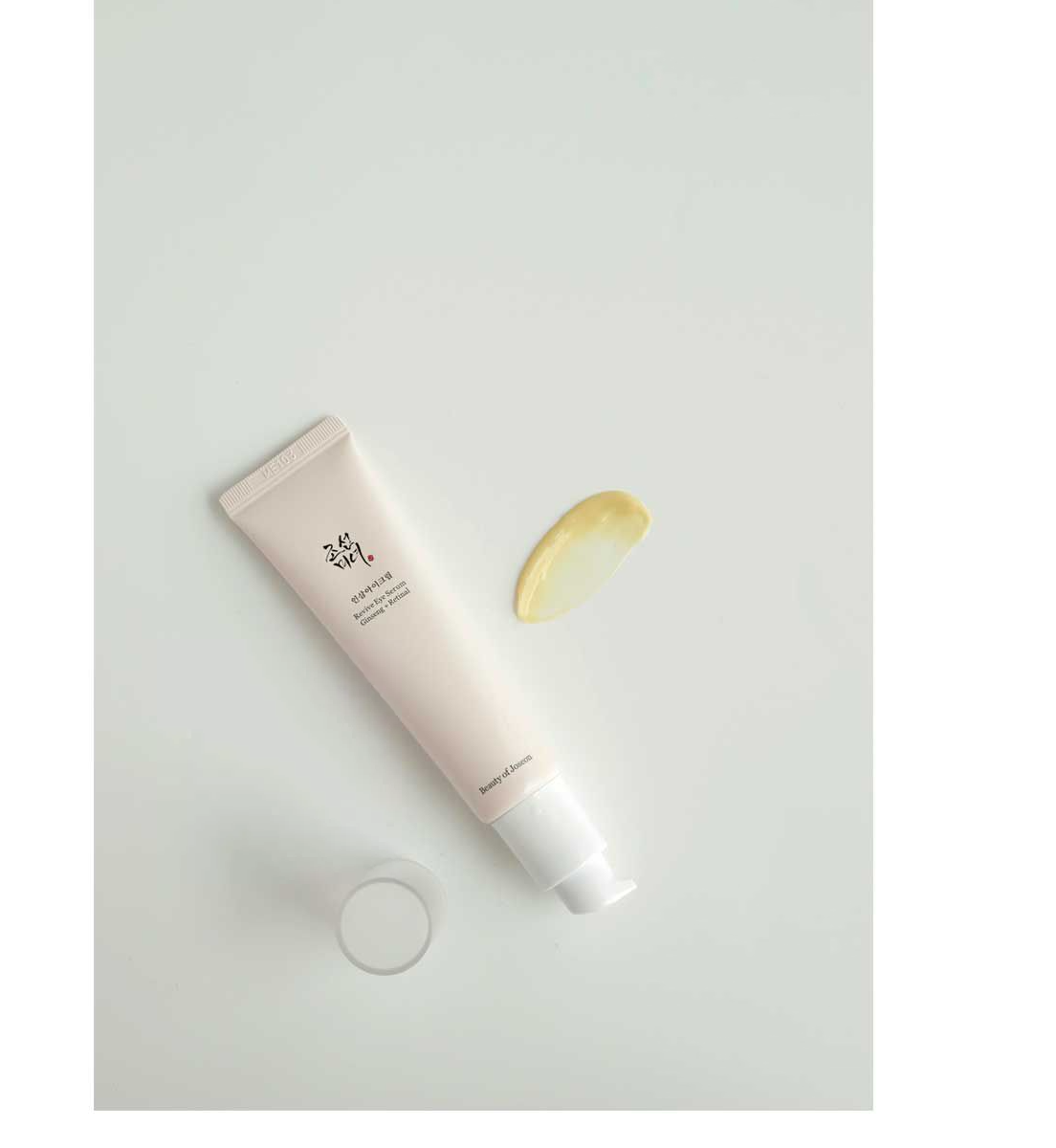 A pink tube of Beauty of Joseons Revive Eye Serum: Ginseng + Retinal rests on a white surface, with a smear revealing its creamy, yellowish texture infused with ginseng for wrinkle reduction.