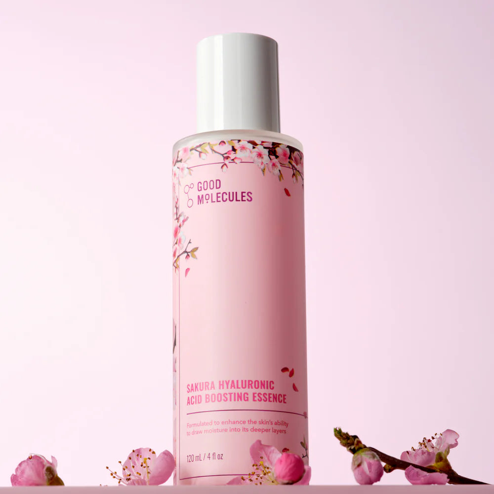 A pink bottle of Good Molecules Sakura Hyaluronic Acid Boosting Essence stands upright among cherry blossoms, emphasizing the hydrating power of sakura flower and hyaluronic acid on a soft pink background.