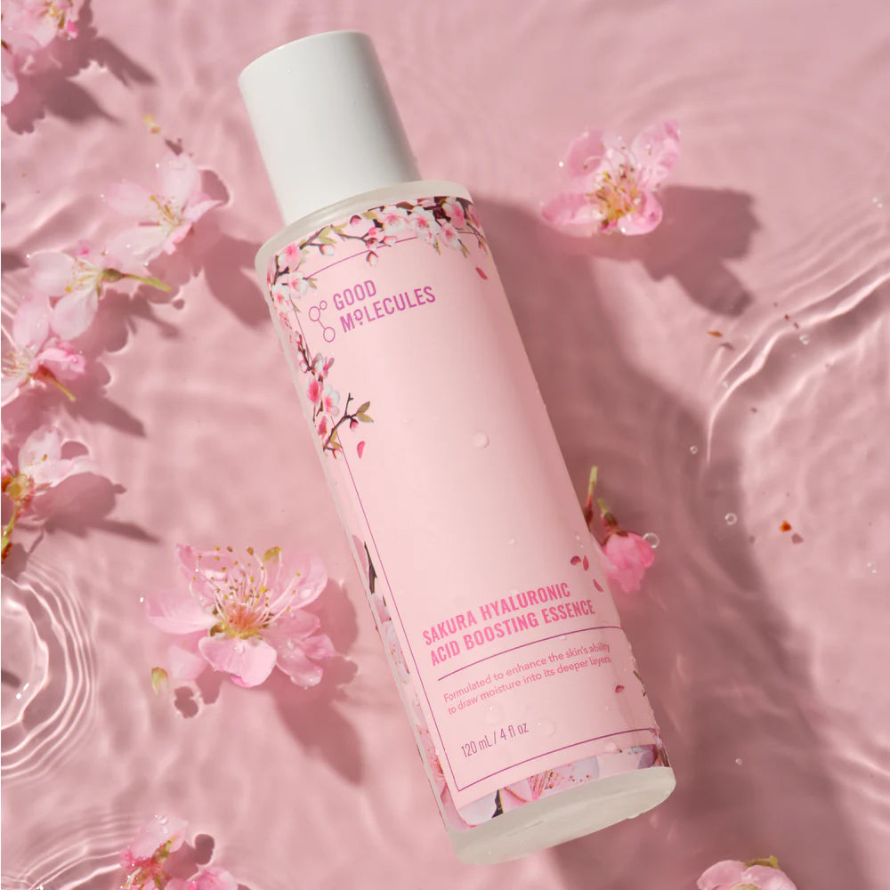 A bottle of Good Molecules Sakura Hyaluronic Acid Boosting Essence floats on pink water amid cherry blossom petals, with floral designs emphasizing its hydrating benefits from hyaluronic acid and sakura flower.