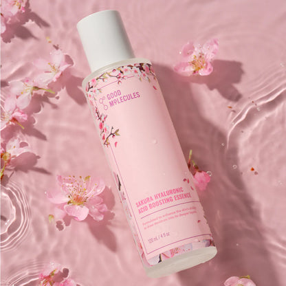 A bottle of Good Molecules Sakura Hyaluronic Acid Boosting Essence floats on pink water amid cherry blossom petals, with floral designs emphasizing its hydrating benefits from hyaluronic acid and sakura flower.