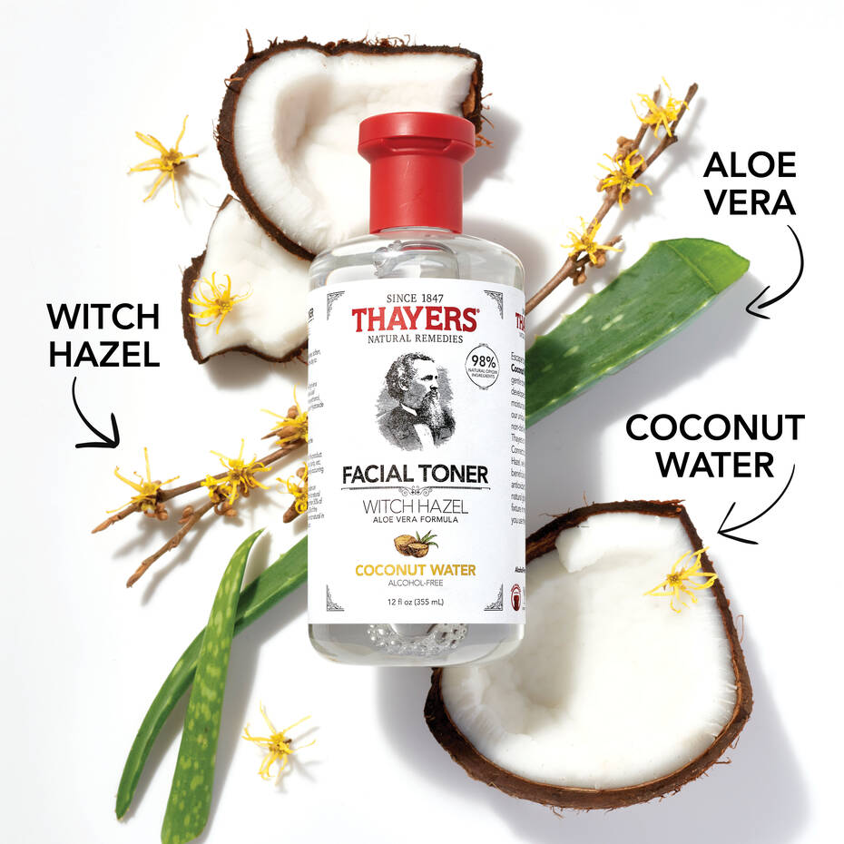 A THAYERS Facial Toner bottle is set among aloe vera leaves, coconut halves, and witch hazel twigs. The label highlights key ingredients: witch hazel, aloe vera, and coconut water. Arrows emphasize the natural elements enhancing this Coconut Water facial toner.