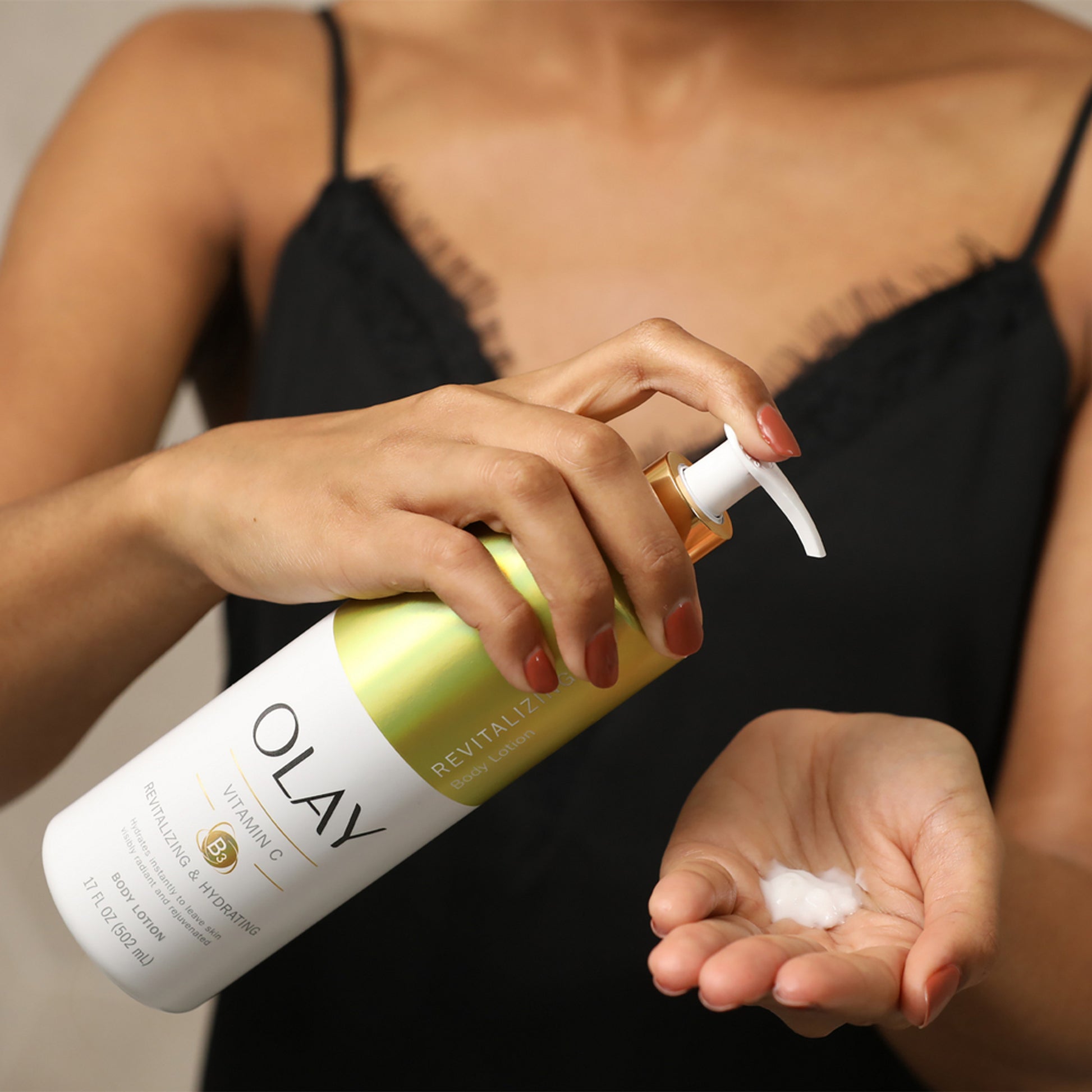 Wearing a black camisole, someone pumps OLAYs 17 fl oz Revitalizing & Hydrating Body Lotion with Vitamin C into their palm for rejuvenating hydration and Vitamin B3 Complex benefits. They hold the bottle in one hand while dispensing lotion with the other.
