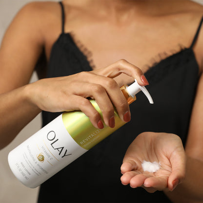 Wearing a black camisole, someone pumps OLAYs 17 fl oz Revitalizing & Hydrating Body Lotion with Vitamin C into their palm for rejuvenating hydration and Vitamin B3 Complex benefits. They hold the bottle in one hand while dispensing lotion with the other.