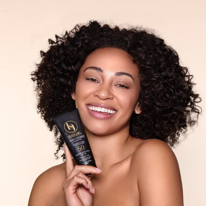 A person with curly hair smiles, holding a black tube of Black Girl Sunscreen Make It Hybrid™ SPF50 - 89ml against a beige background.