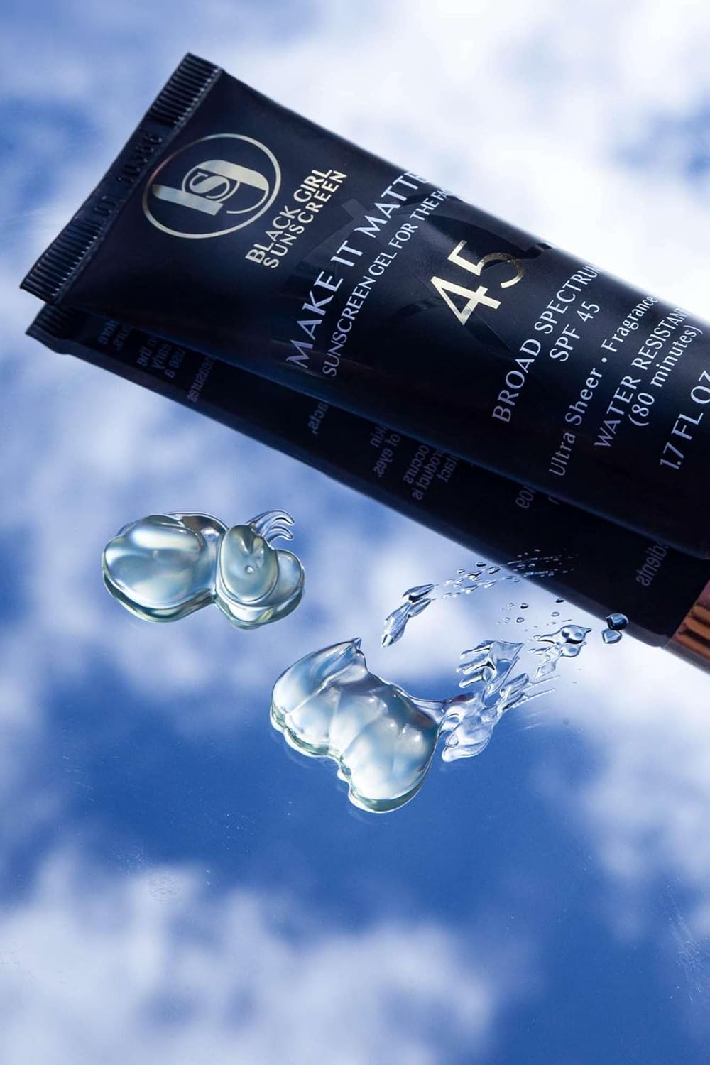 A black tube of Black Girl Sunscreen Make It Matte™ SPF 45 - 50ml is angled against a cloudy sky. Two blobs of vegan sunscreen are shown next to it, capturing light and reflections—ideal for women of color seeking flawless protection.