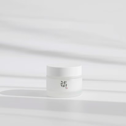 A Beauty of Joseon Dynasty Cream jar with black and red Korean text on the label sits on a smooth light gray surface. Designed for a glass-like complexion, it embodies minimalist elegance. The blurred background features patterns enhancing its allure with Korean ingredients.