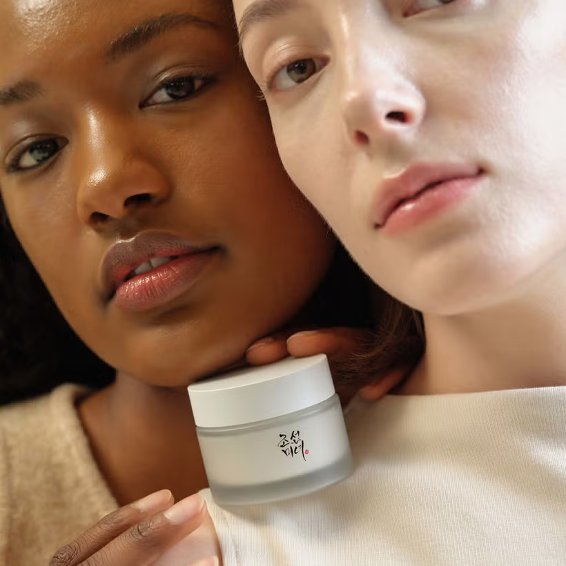 Two women, one with dark skin and the other with light, hold a Beauty of Joseon Dynasty Cream jar. Their radiant complexion highlights the skincare product. One hand rests gently on the others shoulder, symbolizing unity through Korean ingredients.