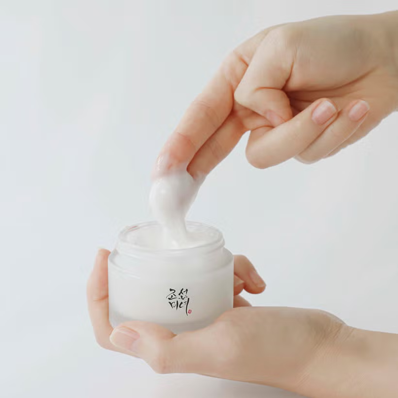A hand gently dips fingers into a jar of Beauty of Joseon Dynasty Cream - 50ml, featuring Korean text and promising a glass-like complexion, set against a minimalist white background.