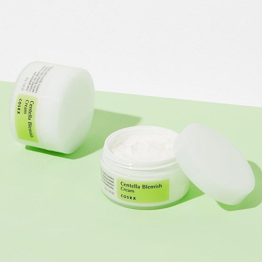 Two jars of COSRX Centella Blemish Cream - 30g, known for treating acne scars with Centella asiatica, are on a light green and white surface. One jar is open, showing the smooth cream inside; the other is closed and tilted. The green labels harmonize with the surface colors.