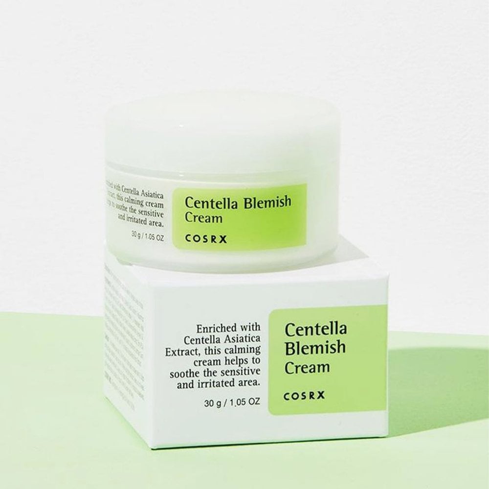 A white jar of COSRX Centella Blemish Cream - 30g is placed on its matching box against a light green and white backdrop, with the box emphasizing Centella Asiatica Extract for its calming effects, ideal as a healing cream for sensitive skin and acne scars.