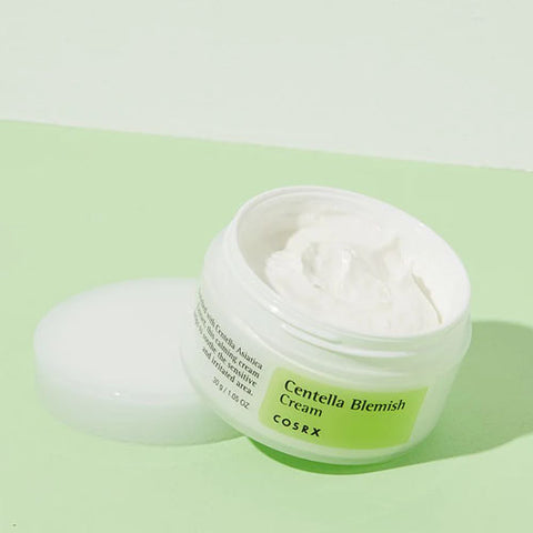 An open white jar of COSRX Centella Blemish Cream - 30g rests on a light green surface, showcasing the soothing cream inside. Enriched with Centella asiatica, this healing cream is perfect for reducing acne scars, with its lid neatly placed beside it.