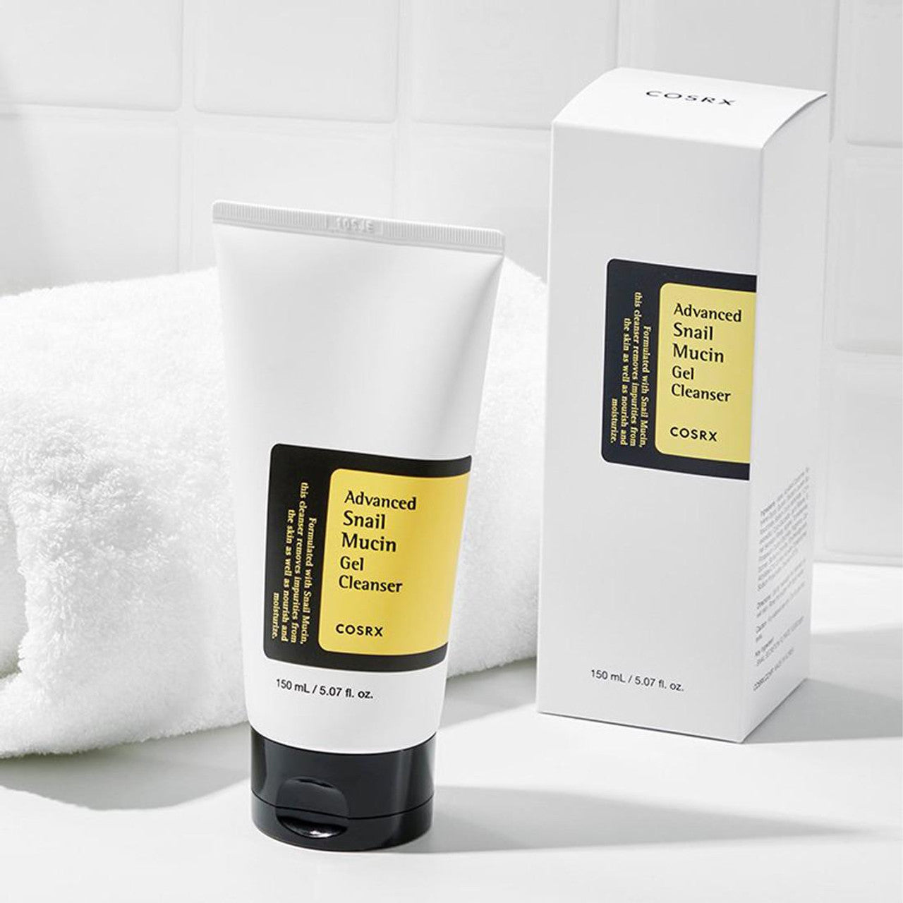 The COSRX Advanced Snail Mucin Gel Cleanser 50ml rests next to its box on the bathroom counter, offering a moisture seal for your skincare routine, while a neatly folded white towel enhances the tranquil atmosphere.