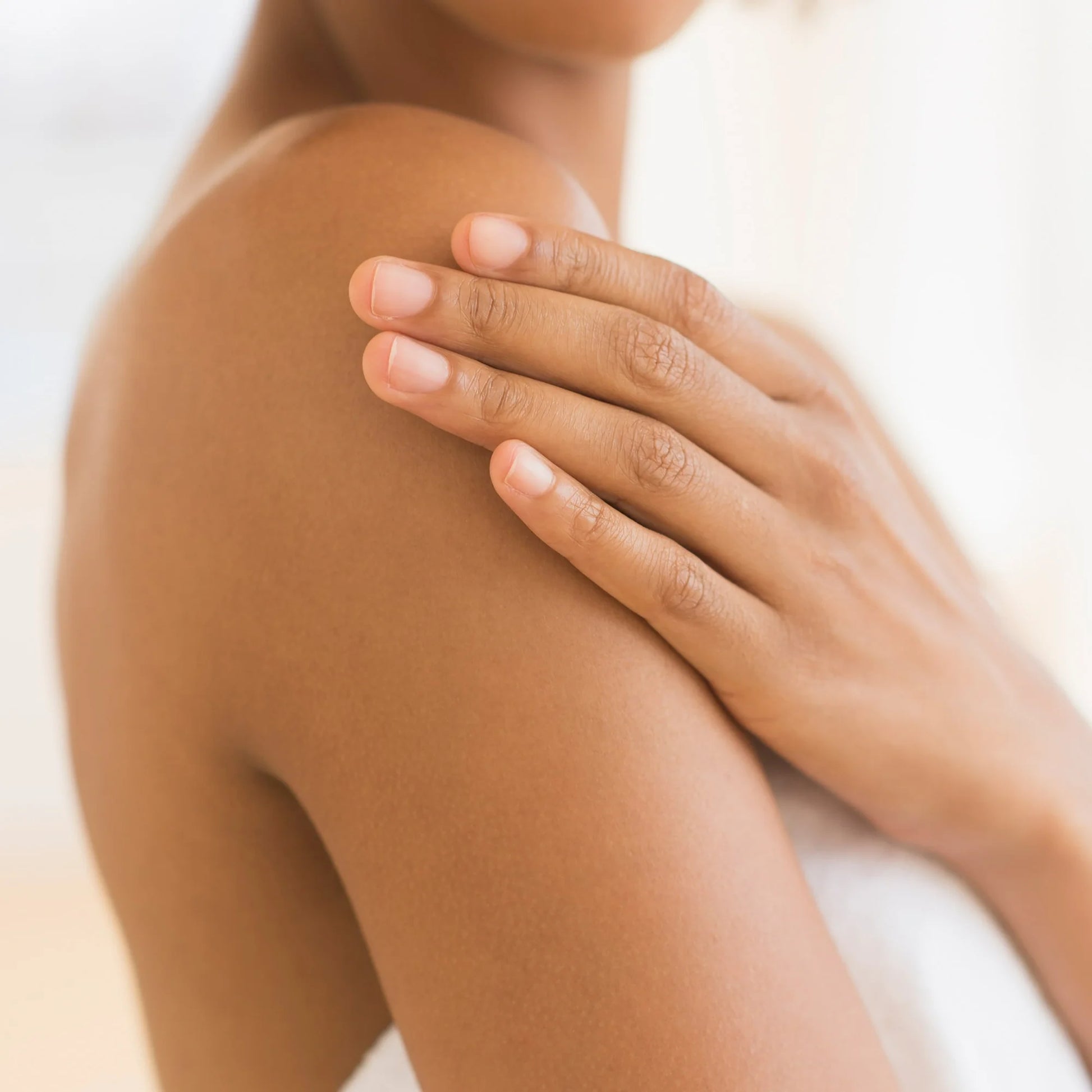 Close-up of a woman's shoulder and hand with smooth, moisturized skin. Her hand gently rests on her shoulder, showcasing healthy and hydrated skin. The background is softly blurred with warm, natural lighting.