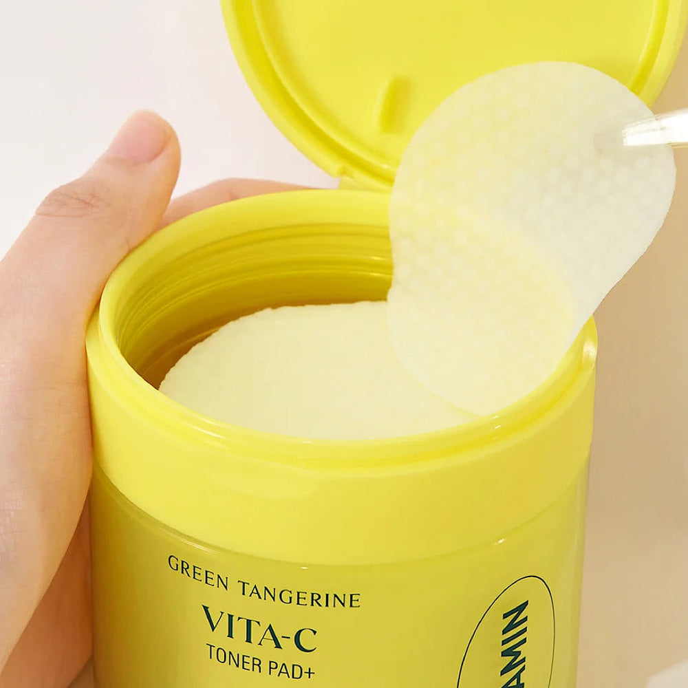 A hand holds a cotton pad from an open Goodal Green Tangerine Vita C Toner Pad container. The lid is open, showing textured circular pads inside that are ideal for addressing dark spots and hyperpigmentation with Vitamin C.