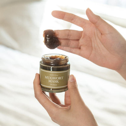 Hands gently cradle a jar of IM FROMs Im From Mugwort Mask 110g, as fingers scoop a bit of the dark brown Korean vegan mask, renowned for soothing sensitive skin, with a green label and white text against a softly blurred background.