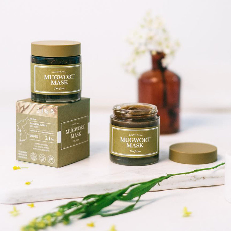 Two jars of Im From Mugwort Mask 110g by IM FROM sit on a marble surface, one open to show the mugwort inside. Theres also a brown glass bottle and a small yellow flower sprig, perfect for soothing sensitive skin.