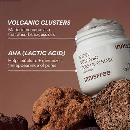 An INNISFREE Super Volcanic Pore Clay Mask 100ml jar rests on volcanic rocks, showcasing its ingredients: volcanic clusters to control sebum and AHA (lactic acid) for exfoliating and minimizing pores, helping with blackhead removal. The background is a warm brown.
