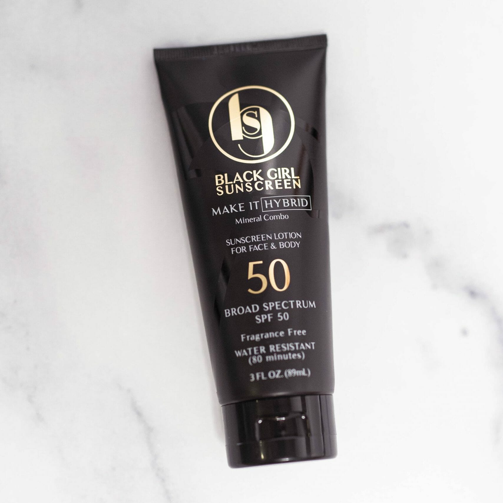 Black Girl Sunscreen Make It Hybrid™ SPF50 in an 89ml tube sits on a white marble surface, featuring sleek black packaging with gold and white text. This broad-spectrum, fragrance-free, water-resistant sunscreen is formulated for face and body using both mineral and synthetic ingredients.