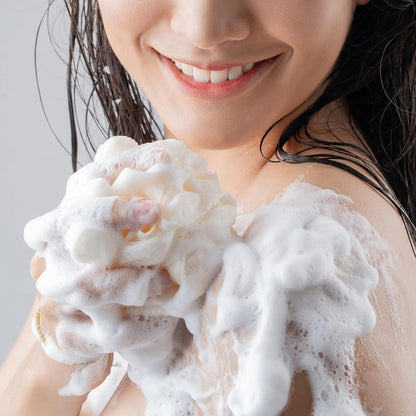 A person with wet hair smiles while applying Olay Cleansing & Renewing Nighttime Body Wash, with Vitamin B3 and Retinol, using a bath sponge. White foamy soap bubbles cover the sponge and part of their skin against a light gray background.