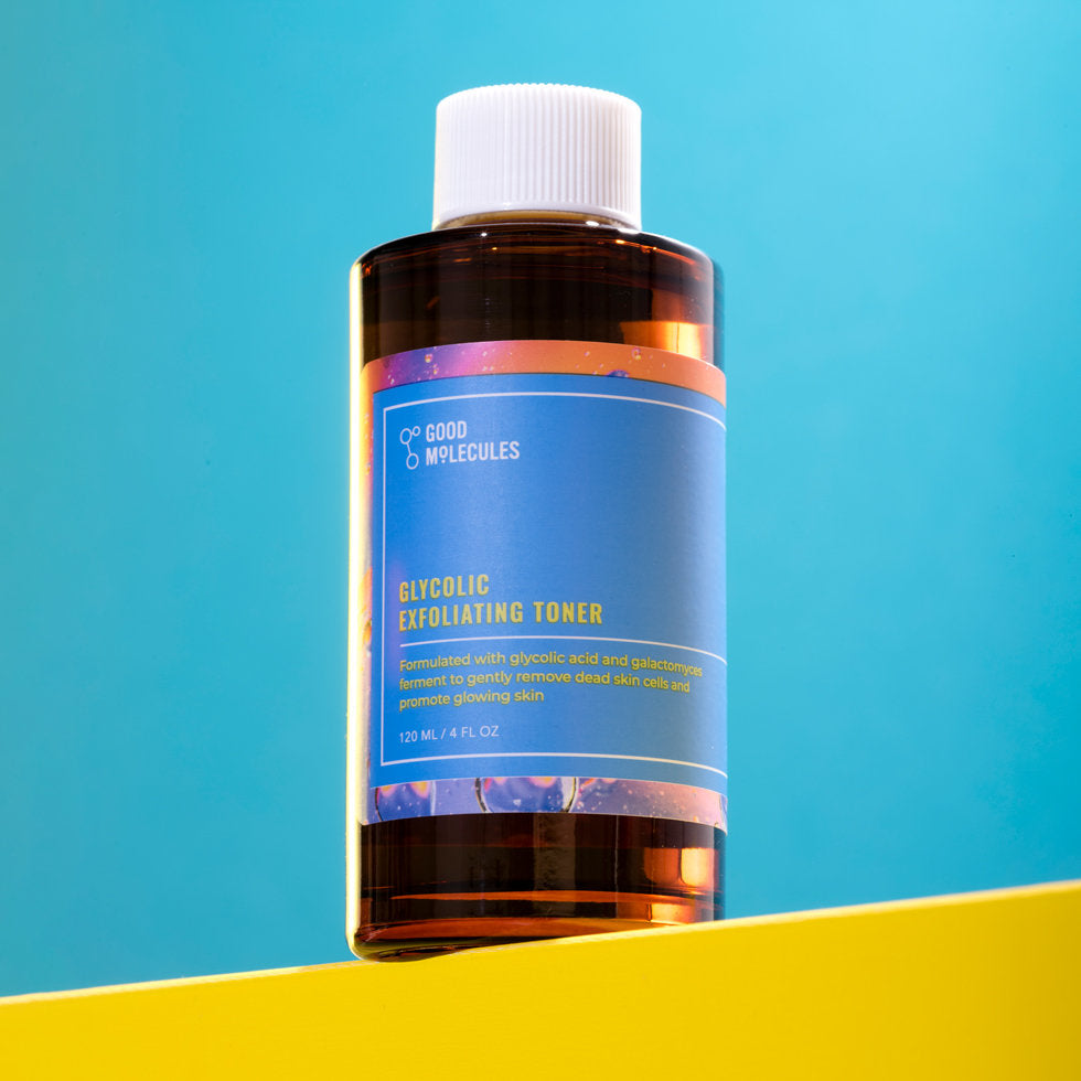 A bottle of Good Molecules Glycolic Exfoliating Toner with a blue label and white cap sits on a yellow surface against a light backdrop. The label promotes its glycolic acid and galactomyces ferment for radiant skin.