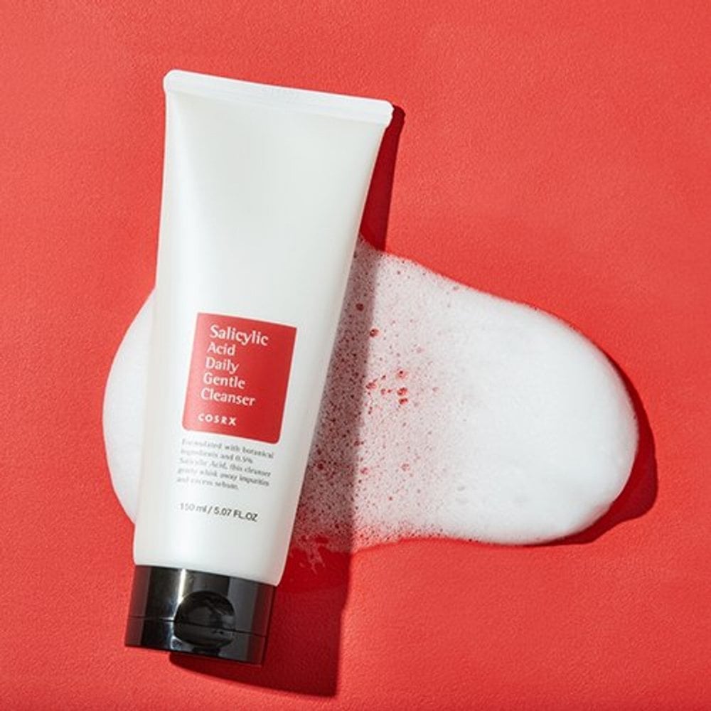 A white tube of COSRX Salicylic Acid Daily Gentle Cleanser 150ml sits on a red surface, showing off its red label and black cap. Beneath it lies a blob of foamy cleanser ideal for acne-prone skin.