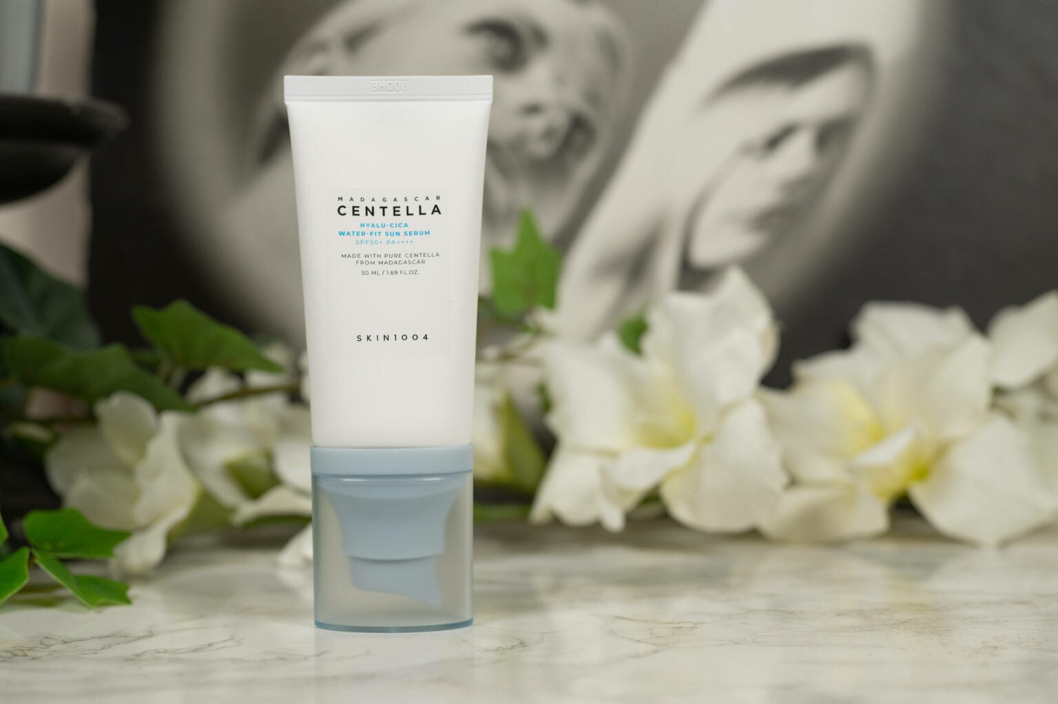SKIN1004 Centella Sun Serum standing on a marble surface with white flowers, highlighting its SPF50+ protection and hydrating formula for all skin types
