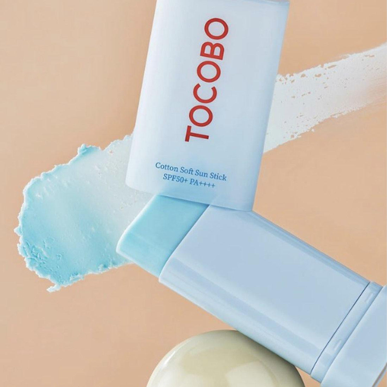Two light blue Tocobo Cotton Soft Sun Sticks (SPF50+ PA++++), featuring red TOCOBO text, are set against a beige background. A matching blue smear extends from the top right, with a beige round object below, emphasizing this 19g vegan sun sticks charm.